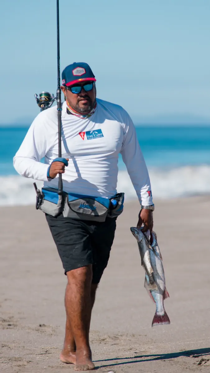 Pescador de orilla caminando por la playa con caña y corvina capturada